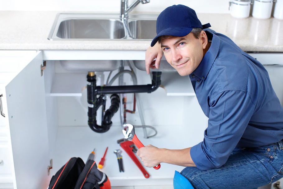 A plumber Toronto in a blue uniform and cap is kneeling by an open kitchen cabinet, holding a wrench and working on pipes under the sink. Various tools are spread out on the floor nearby.