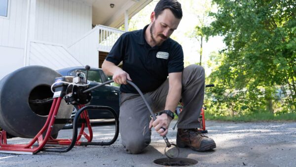 A plumber Toronto kneels on the ground outdoors, using a plumbing snake to clear a drain. Equipment is set up beside him, with a house and trees visible in the background.