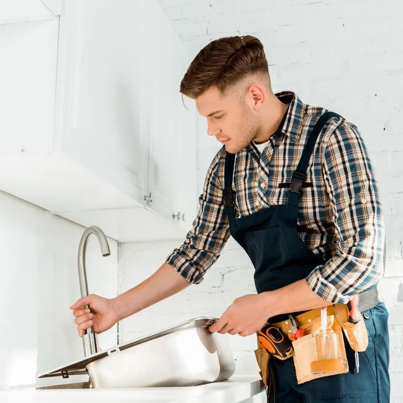 A plumber Toronto wearing a plaid shirt and overalls installs a kitchen sink, holding the faucet with one hand and supporting the sink with the other. Tools are visible in the belt around his waist.