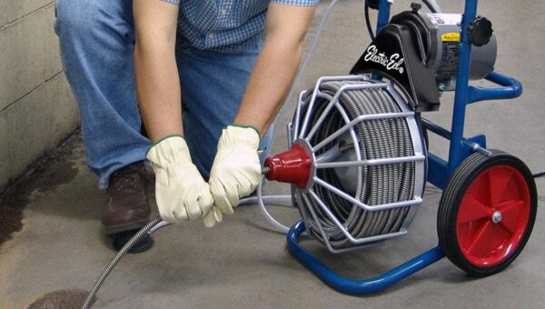 A plumber Toronto wearing gloves and a plaid shirt uses a motorized drain snake machine with a metal coil and red wheels to unclog a floor drain in a concrete surface.