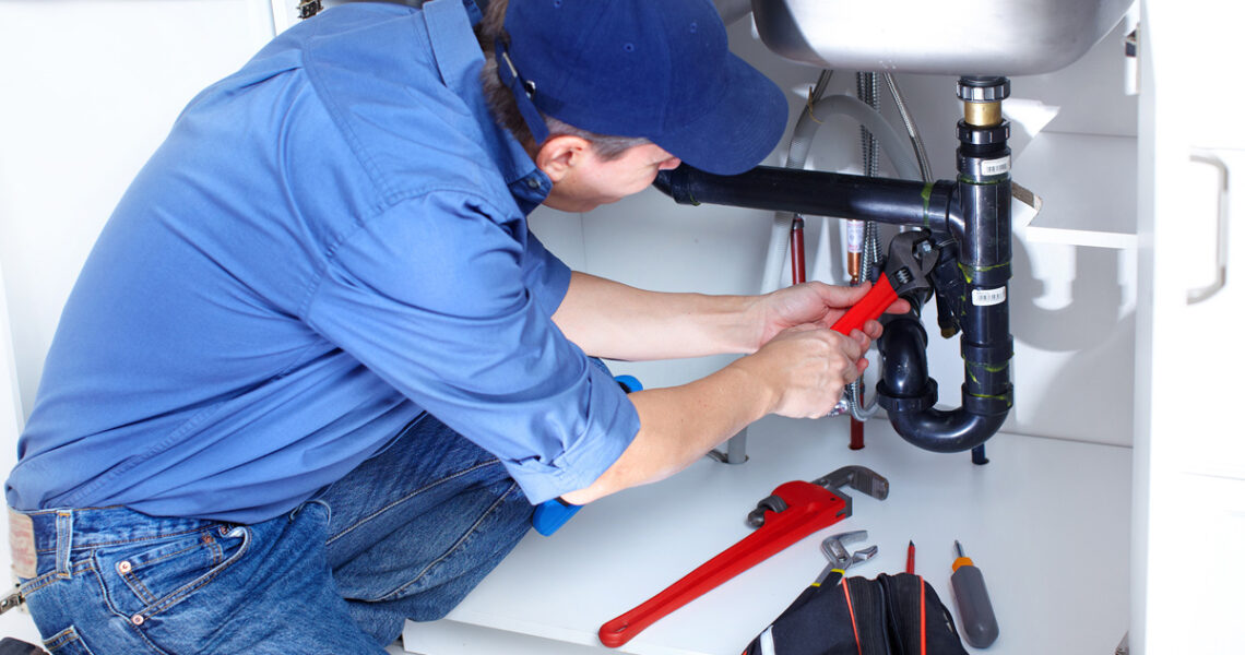 A plumber wearing a blue shirt and cap kneels under a kitchen sink, using a red pipe wrench to fix black pipes. A toolbox with tools lies open on the floor beside him.