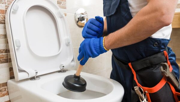 A plumber Toronto professional wearing blue gloves and a tool belt uses a plunger to unclog a white toilet in a bathroom with tiled walls.