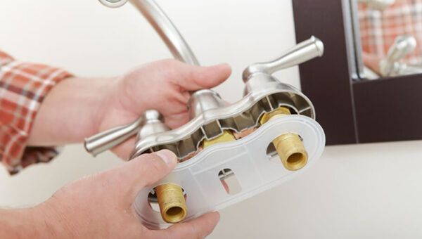 A plumber Toronto in a plaid shirt is installing or repairing a metal bathroom faucet, holding it near a sink with visible water supply connections underneath.