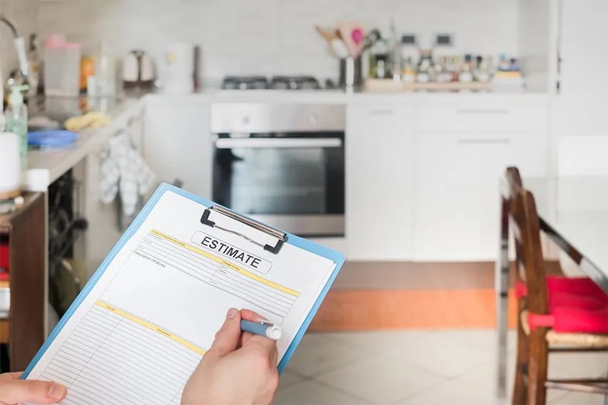 A person holding a clipboard with an estimate form, illustrating how it works while standing in a modern kitchen with a stove, countertop appliances, and dining area in the background.