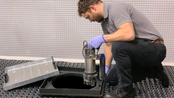 A plumber Toronto kneels on a textured floor, lowering a metallic device into an open rectangular hatch with the removed lid beside it. He appears focused on carefully placing the equipment.