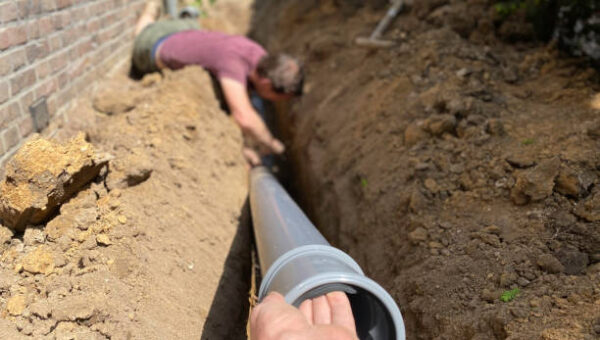 A plumber Toronto places a large PVC pipe into a trench beside a brick wall, while another person in the background helps align the pipe. Soil is piled on both sides of the trench.