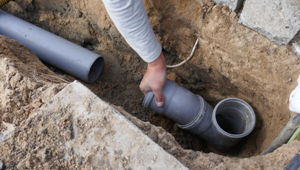 A plumber in Toronto connects two gray PVC pipes in a trench dug into sandy soil, part of an outdoor plumbing or drainage installation project.