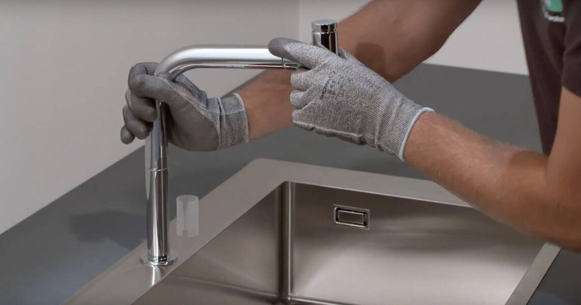 A plumber Toronto is wearing gray gloves while installing or adjusting a modern metal kitchen faucet over a stainless steel sink with a gray countertop.