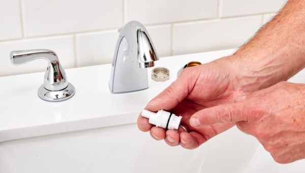 A person holds a faucet cartridge next to a bathroom sink with a disassembled faucet, perhaps preparing for repairs by a plumber Toronto trusts. The countertop and white tiled wall are visible in the background.