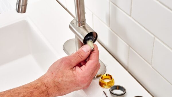 A plumber Toronto is repairing a metal bathroom faucet, removing or installing a cartridge with several faucet parts, including rings and screws, placed on the sink beside it. White tiled wall in the background.