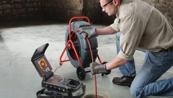 A plumber Toronto wearing safety glasses uses a drain inspection camera, feeding a cable into a floor drain in a basement with concrete flooring and stone walls. The camera monitor displays an image of the pipe interior.