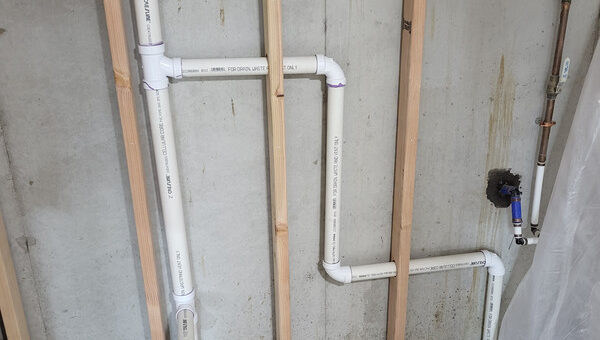 White PVC plumbing pipes are installed between unfinished wooden wall studs against a concrete wall. The floor is partly covered with gravel and debris. Exposed copper plumbing and a plastic sheet are visible, highlighting the need for mold removal Toronto services.