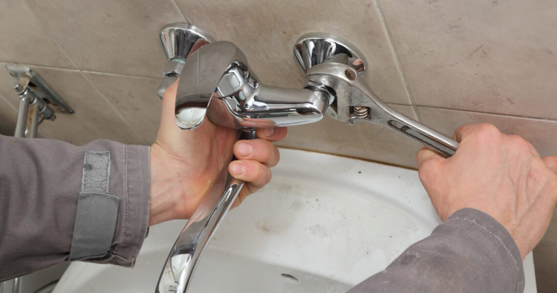 A person uses a wrench to tighten a shiny chrome faucet on a tiled bathroom wall above a white sink.