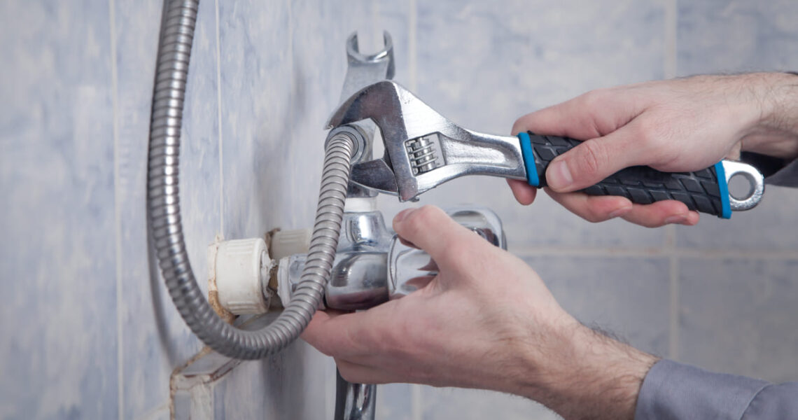 A person uses a wrench to perform shower repair, tightening or loosening a pipe connected to a shower faucet against a tiled bathroom wall.