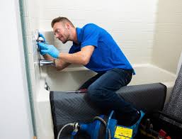 A plumber wearing blue gloves and a blue shirt kneels inside a bathtub, using tools for shower repair on the faucet. A protective mat covers the tub’s edge, with plumbing equipment nearby.
