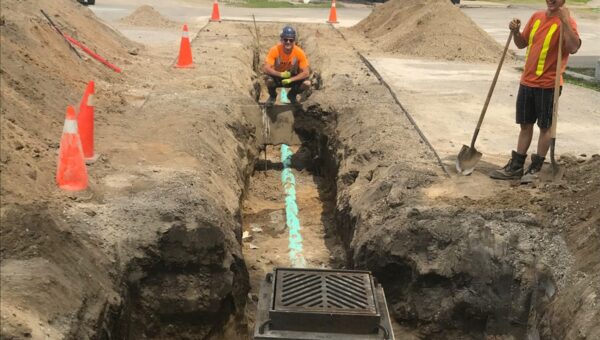Two construction workers in safety vests and helmets work in a deep trench with exposed pipes and a storm drain. Orange cones mark the area, highlighting safety standards similar to those followed by mold removal Toronto services.
