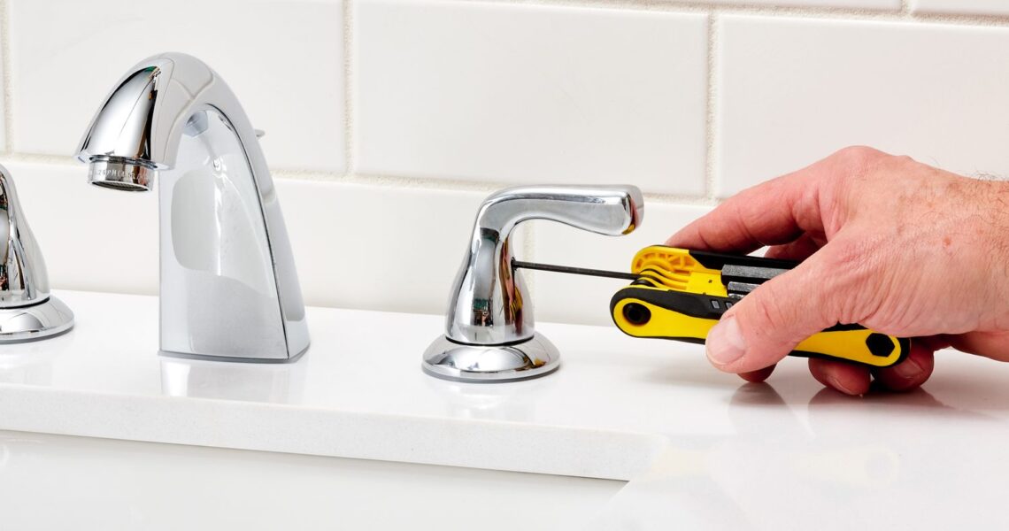 A person uses a yellow-handled hex key wrench for faucet repair, tightening the handle of a chrome bathroom faucet on a white countertop with a white subway tile backsplash in the background.